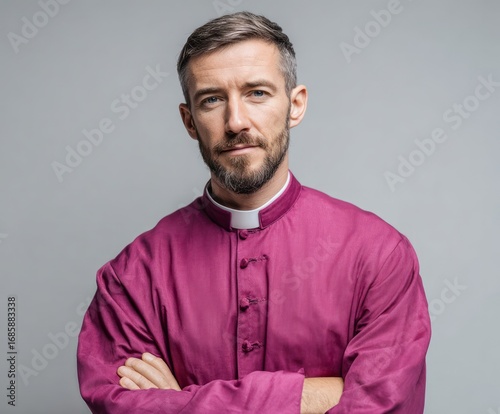 A man in a deep magenta clerical robe, arms crossed, presents a serious and contemplative expression against a neutral backdrop.