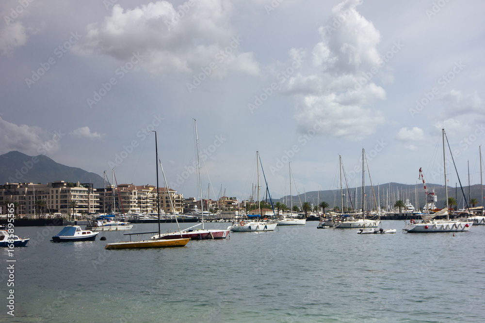 Fototapeta premium Sailboats and yachts moored in harbor with city buildings and mountains, horizontal photo. Tivat Montenegro
