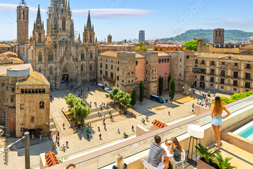 Tourists and young people at a rooftop bar with swimming pool overlooking the plaza and Gothic Quarter across from the Barcelona Cathedral, in Barcelona Spain.