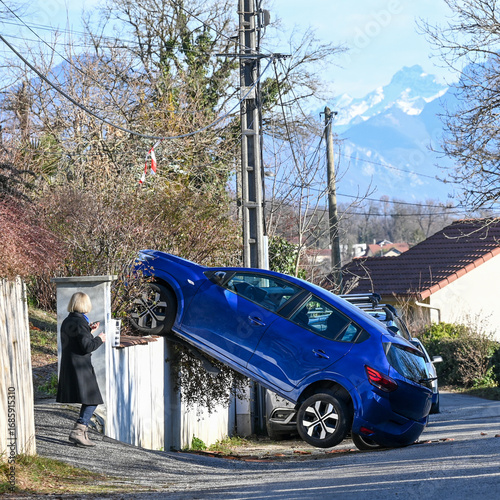 A blue car crashed on top of a fence in a residential area. Unusual accident scene with a woman walking by, mountains in the background, France, French Alps