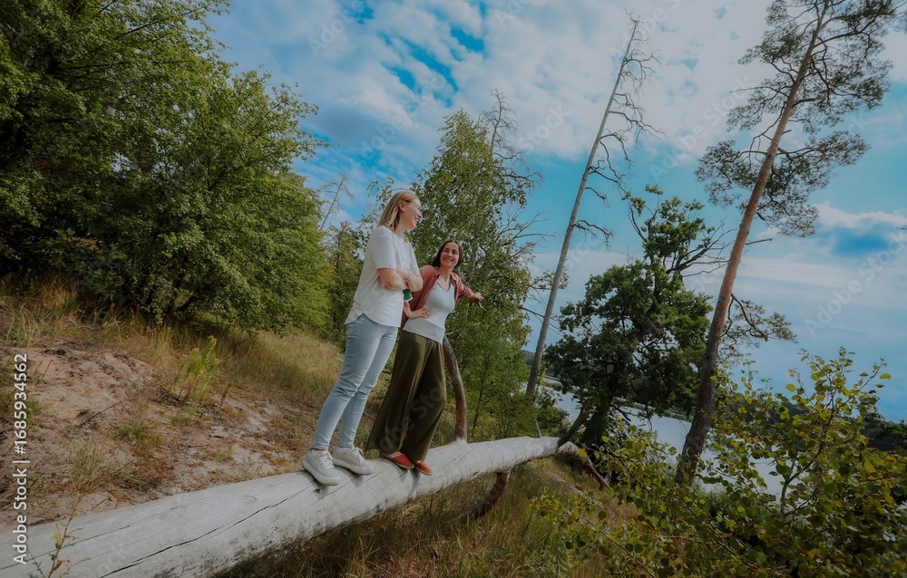 Naklejka premium Two women stand on a fallen tree trunk in a forest near a river, smiling and talking, enjoying nature, friendship, and outdoor adventure under a bright summer sky.