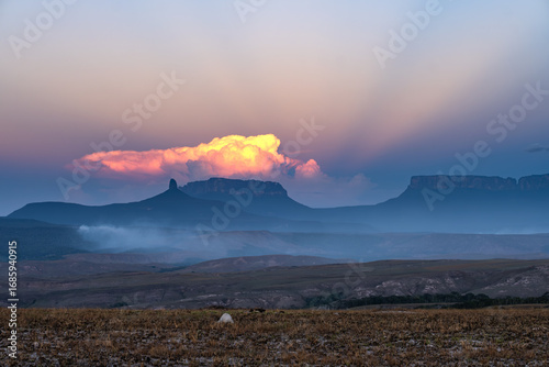 A wide shot of dramatic Venezuelan tepuis at sunset. A vibrant cloud hovers over the iconic flat-topped mountains, highlighting the vast and unique landscape of the Gran Sabana