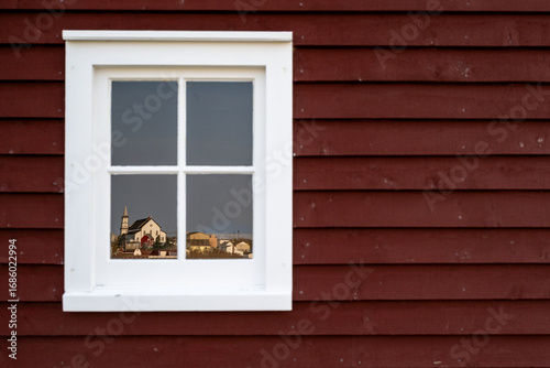 A dark red wooden exterior wall of a house with a four pane glass window. The antique building has wide white trim. The community of Bonavista, a church and house, is reflected in the mirrored glass