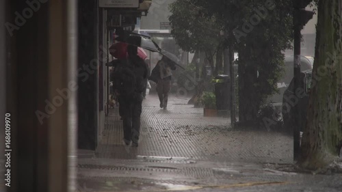 Torrential rain in Buenos Aires, Argentina. Daytime city scene with heavy rainfall, people under umbrellas, cars on wet streets, water puddles splashing, trees, buildings, and urban life. 4K 10-bit fo