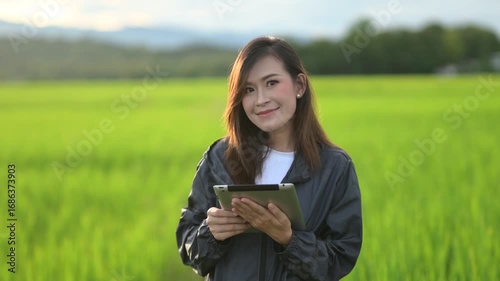 A smiling woman in a grey jacket holds a tablet in a lush green field, radiating positivity and connection with nature.