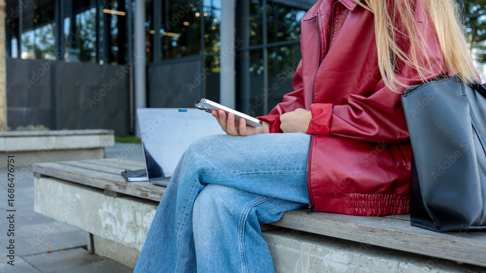 Obraz premium Woman checking her phone outdoors with a laptop nearby.