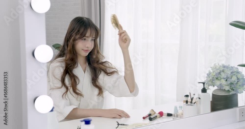 Asian teenage girl in bathrobe brushes wavy hair while smiling at vanity mirror during self care routine in cozy bedroom, expressing joy and natural beauty in relaxed lifestyle setting