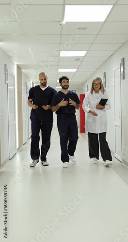 Three healthcare professionals walk and discuss in a hospital hallway, two male doctors in scrubs and a female doctor in a lab coat are talking.