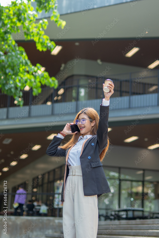 Fototapeta premium Asian businesswoman in suit using mobile phone while standing in front of modern business building.