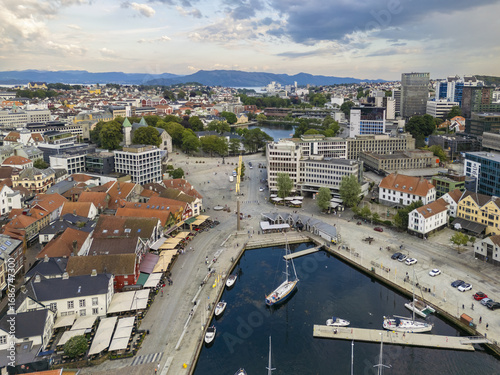 Aerial view of terracotta rooftops cascade towards the shimmering harbor where masts dance against the deep blue water, shadowed by the city skyline, Stavanger, Rogaland, Norway.