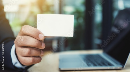 Hand holding blank card with laptop in background on a wooden surface.