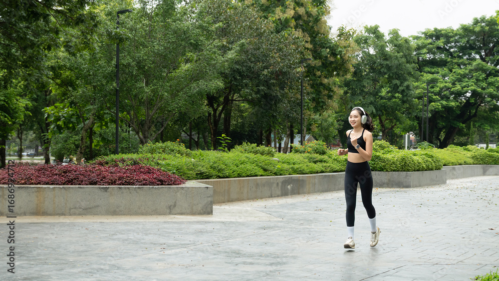 Fototapeta premium Happy woman listening music on earphones while running in nature in the morning. Healthy fitness woman jogging outdoors.