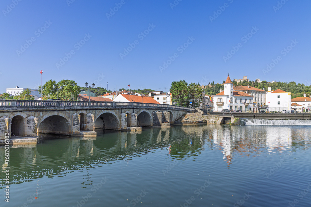Obraz premium An ancient stone bridge with multiple arches spans the Nabão River in Tomar. The town's historic buildings and the castle on the hill are reflected in the calm water.