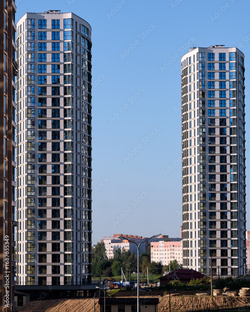 Obraz premium Twin apartment buildings, with repeating window patterns and light gray exteriors, rise above the city landscape under a bright, cloudless blue sky