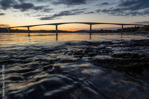 Fotografi Bridge over troubled waters – a picturesque coastline with rocky shore, seaweed, and golden sky during evening light