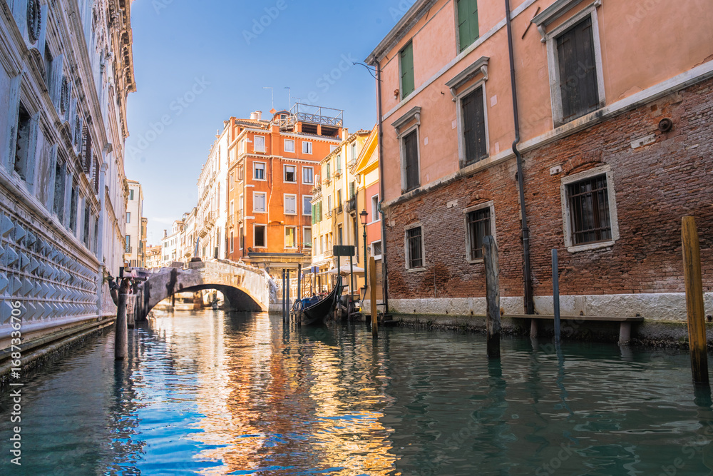 Obraz premium Venetian Canal Scene: Reflections of colorful buildings along a calm canal with a gondola and a small bridge.