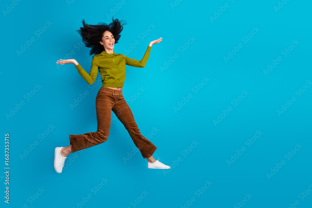 Fototapeta premium Enthusiastic young woman jumps against blue background, showcasing vibrant energy and trendy style, perfect for promoting positivity