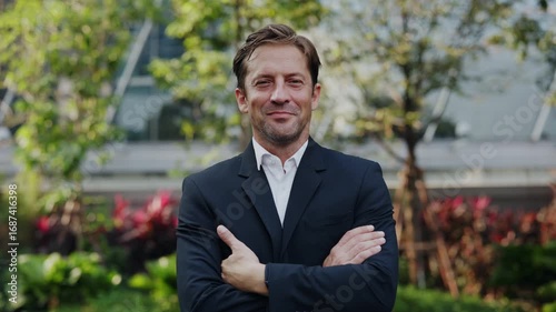 Portrait of happy businessman in suit crosses arms standing in urban park. Mature man entrepreneur smiles conveying success and professionalism in downtown district slow motion