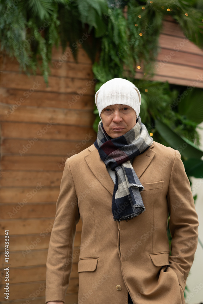 Naklejka premium Winter portrait of caucasian man wearing white beanie and cozy scarf, with blurred Christmas festive background. Cold winter weather. Outdoors. Xmas holiday.