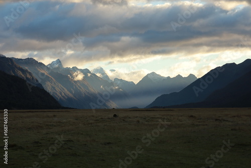 Dramatic light pierces through clouds, illuminating snow-capped mountains, golden plains of Eglinton Valley, trip to Milford Sound, New Zealand, on May 27, 2025. A majestic Lord of the Rings landscape