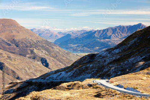May 18, 2025: Crown Range Summit, New Zealand. Winding alpine road through rugged mountains with Queenstown and its airport visible in the stunning Wakatipu Basin. Scenic winter/autumn landscape.