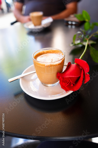 Glass of cortado coffee with red rose on saucer and spoon on a black table