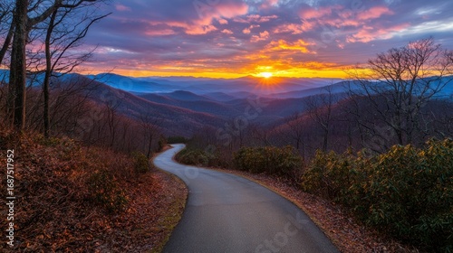 A winding road leads towards a vibrant sunset over layered mountains, framed by trees in late autumn.
