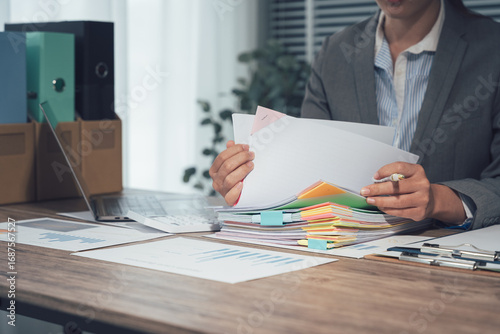 Businessman working through pile of documents to find unfinished documents, information on pile of documents on desk and checking financial documents amidst busy workload.