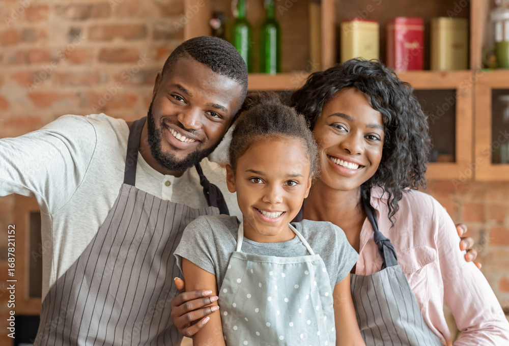 Obraz premium Happy african family taking selfie at kitchen, wearing aprons and smiling at camera