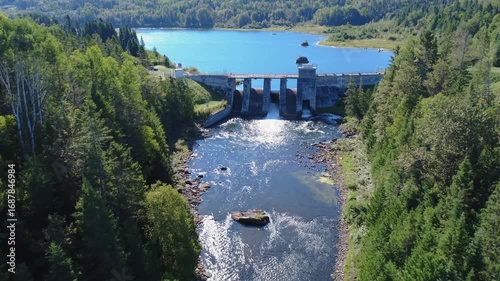 Panoramic aerial view looking down towards a lake and an old hydroelectric dam, passing inside its concrete structure. Mitis Dam, Lake & Mitis River, Quebec, Canada, 2025.