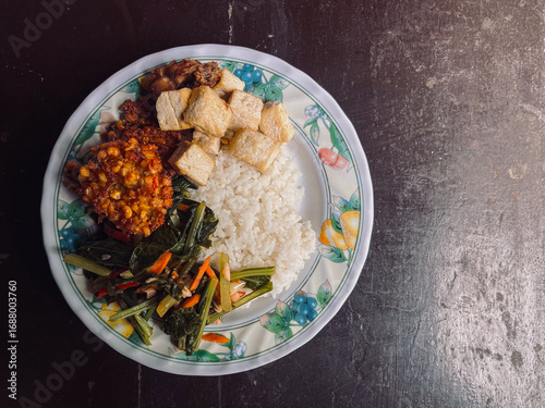 Top view of Indonesian Food with side dishes of mix vegetables, tofu, frikadel or corn fritters, fried chicken, and steamed rice as main food on white plate. Isolated by brown wooden
