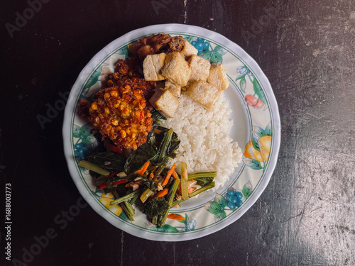 Top view of Indonesian Food with side dishes of mix vegetables, tofu, frikadel or corn fritters, fried chicken, and steamed rice as main food on white plate. Isolated by brown wooden