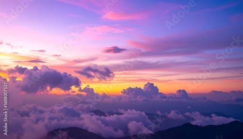 A stunning vista of a mountainous landscape at sunset, the sky ablaze with vibrant hues of purple, orange, and pink. The clouds are fluffy, floating above the peaks, casting a mystical atmosphere
