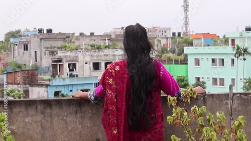 Back view of a beautiful Bengali woman in a maroon saree with long black hair, standing alone on a rooftop and enjoying the fresh air while looking out at the urban cityscape.