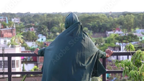 A serene Bangladeshi Muslim woman in a green abaya and khimar stands on a balcony, her clothes swaying in the breeze as she peacefully observes the outdoor beauty.