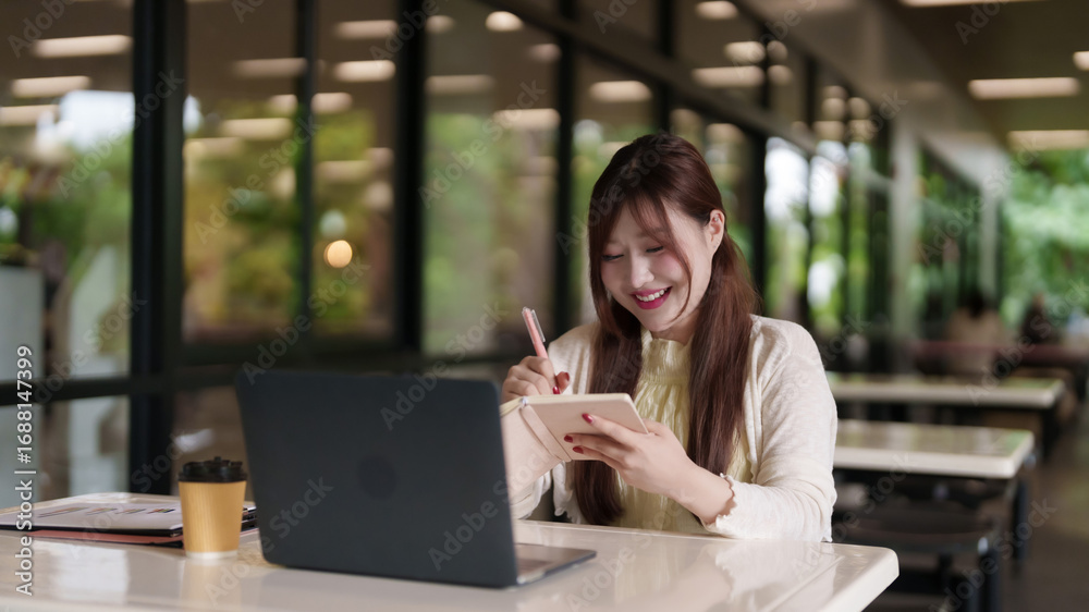 Fototapeta premium Adult Asian freelance professional works productive outdoor cafe writing notes laptop computer natural light happy lifestyle
