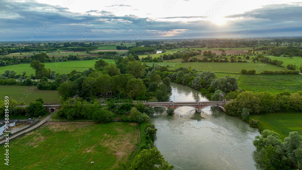 Fototapeta premium Sunset aerial landscape showcasing Oglio River winding near Pontevico, Italy, revealing verdant countryside, infrastructure, agricultural fields, and distant industrial structures
