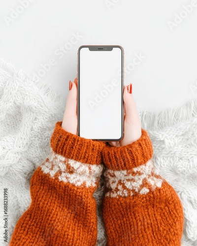 Hands holding a phone, wearing an orange sweater, on a white blanket