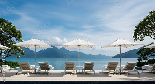 High-angle view of empty lounge chairs and white umbrellas by a tranquil turquoise ocean, conveying a serene and peaceful mood, with mountains in the background; a