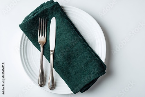 A dark teal linen napkin sits on a white plate, alongside a fork and knife, on a white background