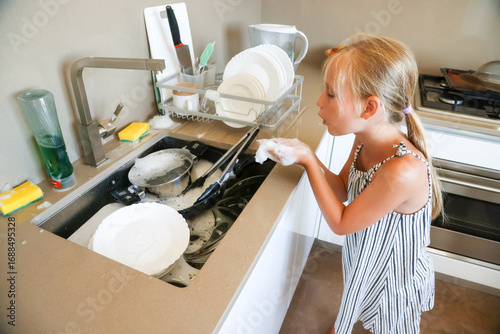 Little girl blowing soap foam bubbles while helping wash dishes in modern home kitchen