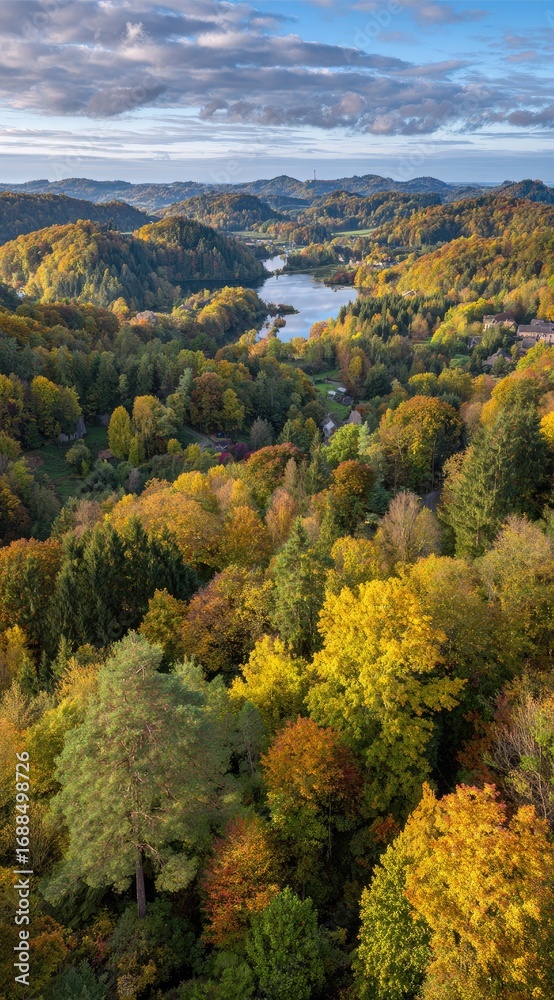 Fototapeta premium An aerial view of an autumn landscape featuring lush forests in warm colors, rolling hills, and a tranquil lake under a sky streaked with clouds