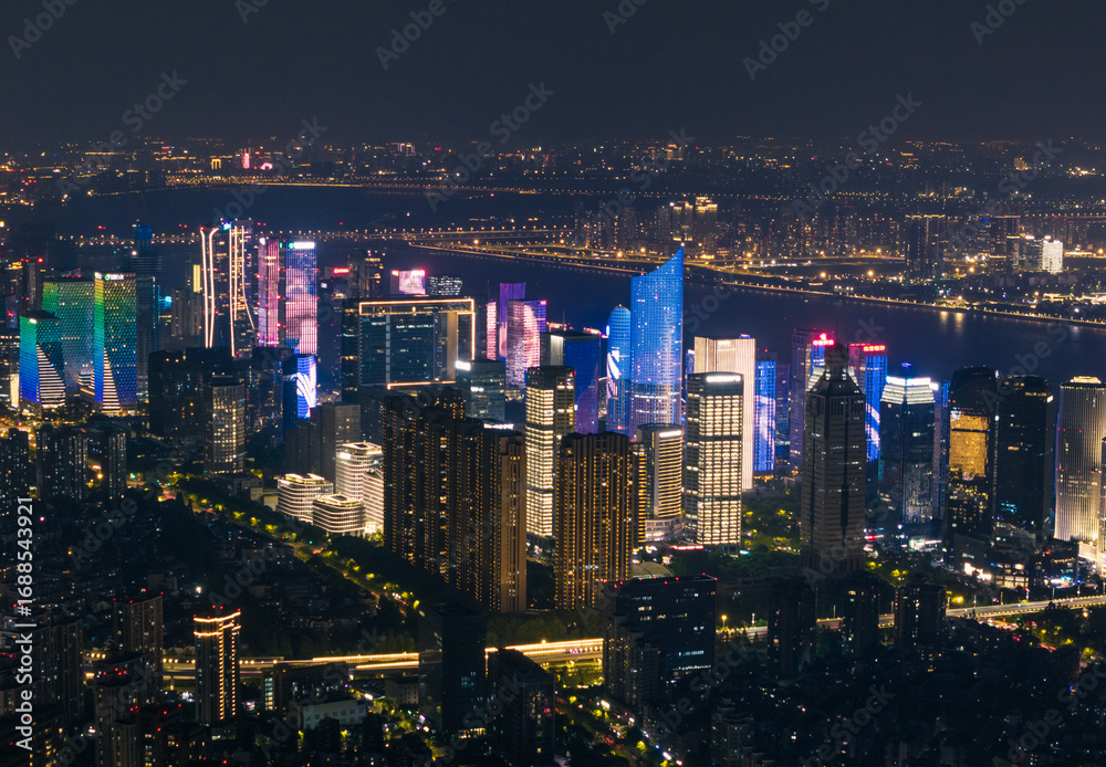 Fototapeta premium Distant cityscape view of lit skyscrapers and buildings in Hangzhou, China at night with illuminated river in the background