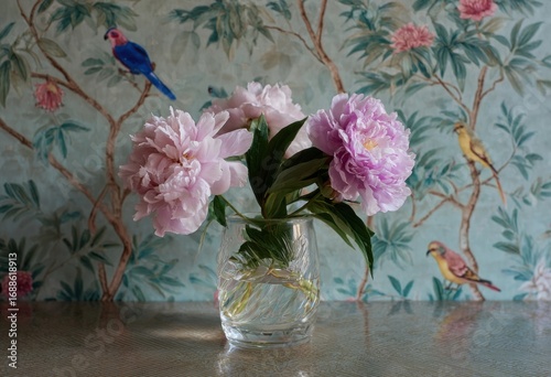 Pink peonies in a glass vase,  against a wallpaper backdrop of birds and flowers