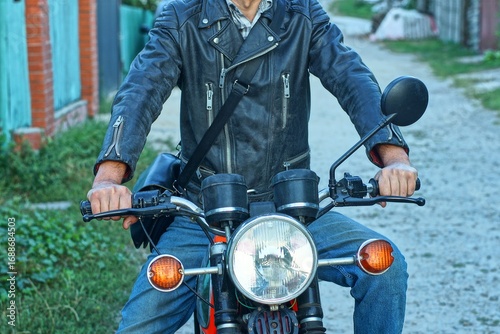 one biker in black old worn with shiny iron zippers and buttons leather stylish fastened with belt and buckle biker jacket and blue jeans sits on motorcycle and holds steering wheel with hands outside