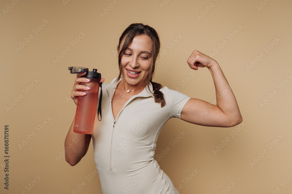 Obraz premium portrait of fit young woman flexes her muscles while holding a water bottle, embodying strength and enthusiasm for fitness on beige background