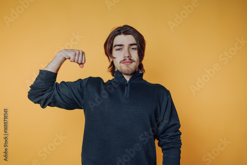 portrait of young man confidently flexes his arm with a playful smile, emphasizing strength and vitality on yellow background