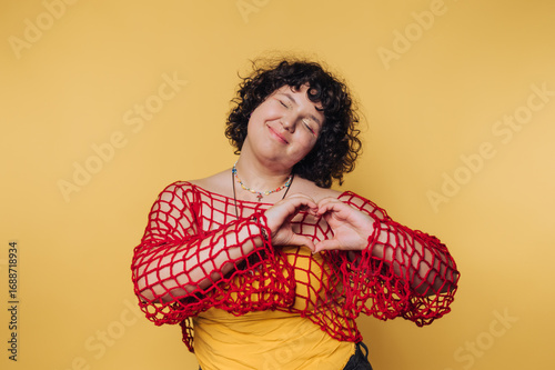 portrait of young woman smiles brightly while forming a heart shape with her hands, symbolizing love and joy in a warm, engaging atmosphere on yellow background