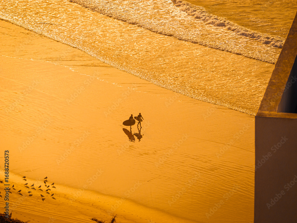 Obraz premium SILHOUETTE: Two surfers walk with their surfboards on wet, golden sand of Moroccan beach. Powerful light of setting sun illuminates the breaking waves, with a flock of small birds scattered nearby.