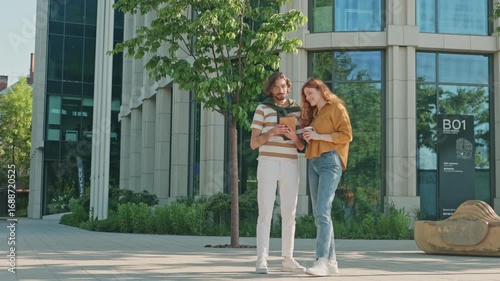 Freelance workers standing outside office center discussing details of project using modern technology and gadgets, while African-American man rides electric scooter down city street.